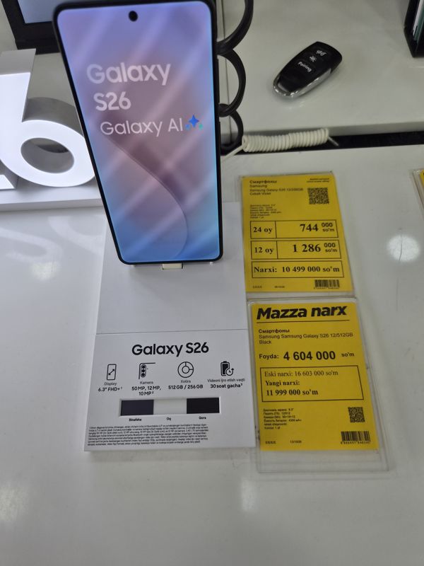 Смартфон Samsung Galaxy S26 на подставке с карточкой характеристик, над ней два жёлтых ценника: Galaxy S26 12/256GB Cobalt Violet (без акции) и Galaxy S26 12/512GB Black («Mazza narx»)