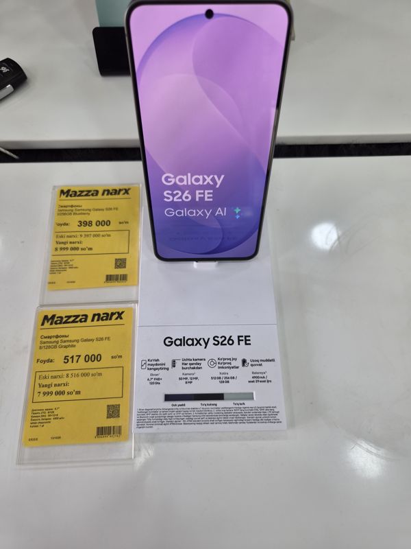 Смартфон Samsung Galaxy S26 FE на подставке с карточкой характеристик, под ней два жёлтых ценника «Mazza narx» один под другим: Blueberry и Graphite