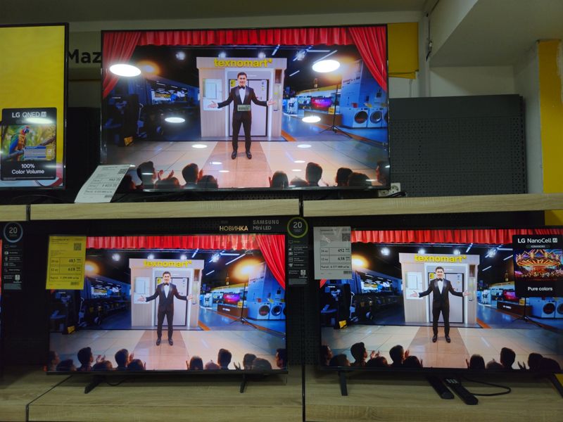 Три телевизора на двух полках: сверху Samsung 51"-60" (ценник 8 355 000), слева внизу Samsung 40"-43" (ценник 5 299 000), справа внизу LG 43NANO80 (ценник 6 377 000); на экранах рекламный ролик магазина