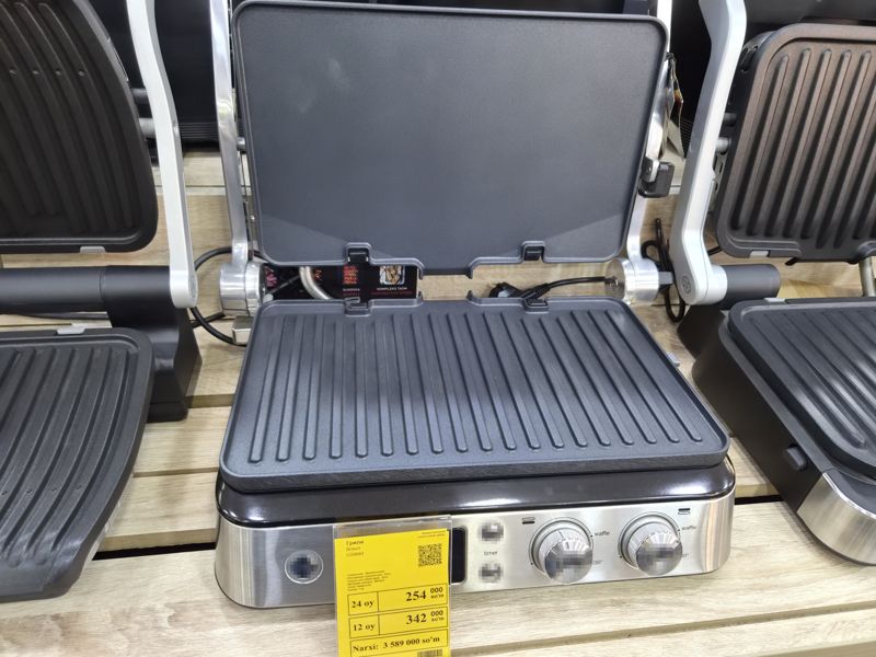 Электрогриль Braun CG9043 с открытой верхней панелью на деревянной полке, спереди жёлтый ценник