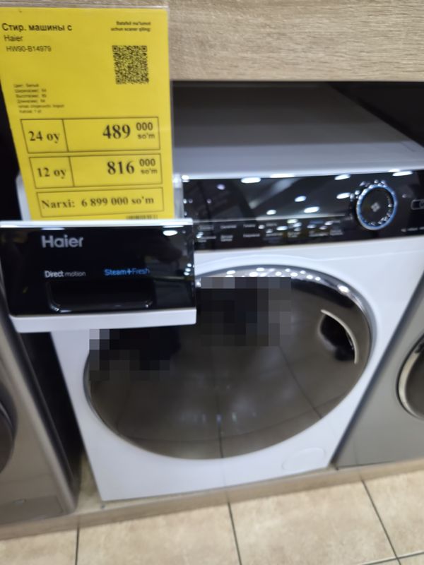 Белая стиральная машина Haier HW90-B14979 с чёрной панелью на стенде, слева жёлтый ценник Техномарта; фото смазано