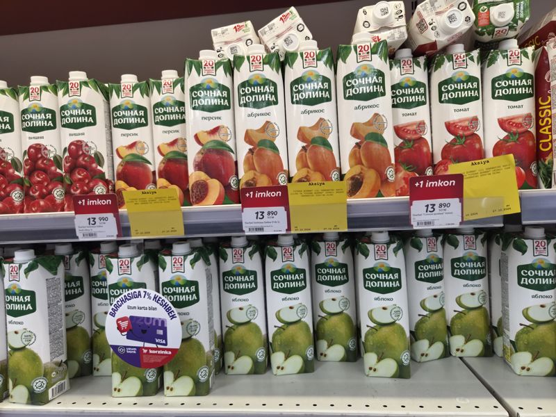 Полка с соками Сочная Долина 1 л в Korzinka (вишня, персик, абрикос, томат, яблоко) с красными плашками «+1 imkon» и жёлтыми карточками «Aksiya»