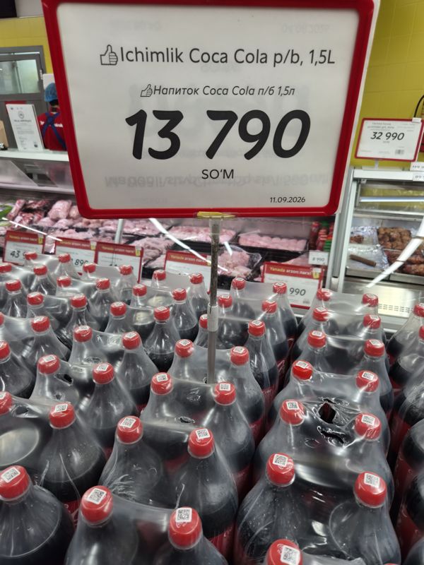 Горка пластиковых бутылок Coca-Cola 1,5 л с крупным ценником 13 790 сум; за ней мясная витрина и жёлтые ценники «Yanada arzon narx!»