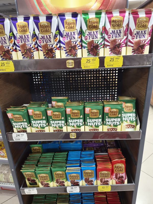 Стойка шоколада Alpen Gold в Korzinka: Max Fun на верхней полке с жёлтыми акционными ценниками, Super Nuts, плитки 80 г внизу