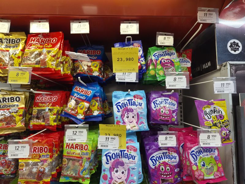Подвесная выкладка жевательного мармелада в Korzinka: Haribo (Happy Cola, Starmix, Worms, Cola Zourr) и Бон Пари (Страна единорогов, Кошмарики, Кислые червячки, Машинки, Кислые лапки), жёлтые карточки «Aksiya 2 + 1»