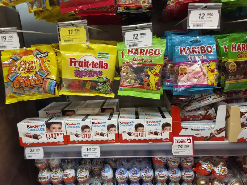 Подвесной мармелад (Chupa Chups, Fruit-tella, Haribo) над полкой Kinder Chocolate и Kinder Bueno с красной плашкой «Narxiga javob»