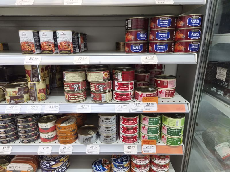 Холодильная витрина с мясными и рыбными консервами в Korzinka: тушёнка из говядины, конины, курицы, шпроты, килька, плов; белые и оранжевые ценники