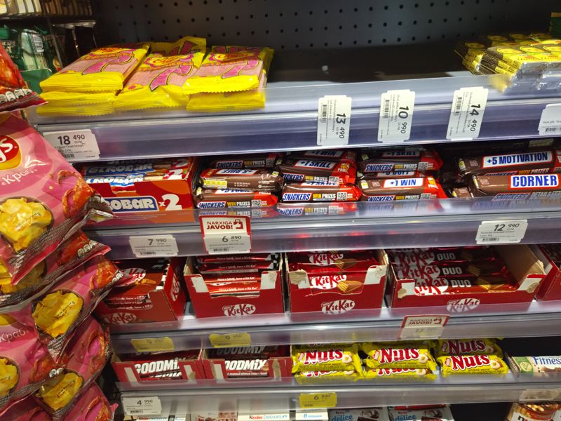 Полки шоколадных батончиков и плиток в Korzinka: Snickers, Strobar, KitKat, Goodmix, Nuts, Chupa Chups Strayps; вертикальные ценники Hershey's, плашки «Narxiga javob!»