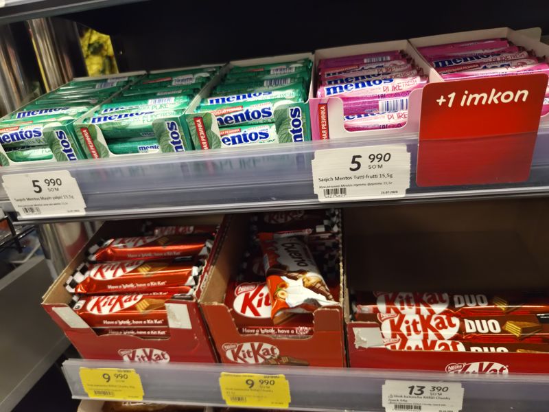 Полки жевательной резинки Mentos и батончиков KitKat в Korzinka; жёлтые ценники KitKat Chunky, красная вставка «+1 imkon»