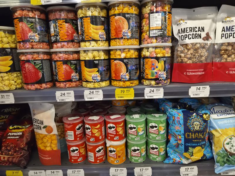 Полки с попкорном Real American (банки), Bisyor American popcorn, Crash, чипсами Pringles в банках и Chaka; ниже ценники