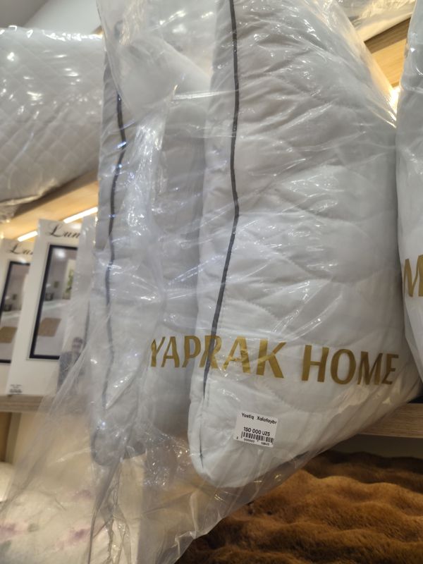 Подушка Yaprak Home в прозрачном чехле, на чехле ценник; рядом другие подушки и коробки Lun...