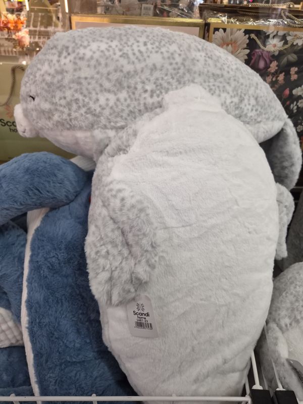 Большая серая мягкая игрушка-тюлень с белым брюхом в проволочной корзине, на боку картонная бирка Scandi home, рядом синяя плюшевая игрушка