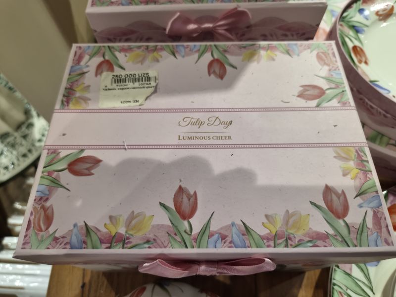 Розовая подарочная коробка с лентой и тюльпанами «Tulip Day, Luminous Cheer» с оторванной наклейкой цены в углу крышки, вокруг другие коробки и тарелки с тюльпанами