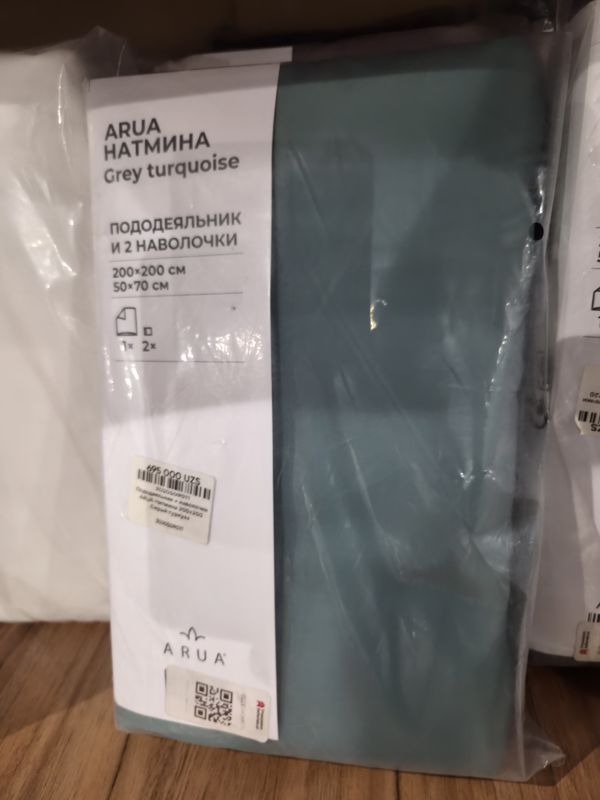 Комплект постельного белья ARUA Натмина серо-бирюзового цвета в прозрачной упаковке на деревянной полке, на упаковке наклейка с ценой и QR, справа соседние упаковки