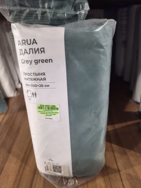 Простыня на резинке ARUA Далия серо-зелёная 160×200×26 см в прозрачной упаковке на деревянной полке, на упаковке наклейка с ценой, выделенная зелёным маркером, и QR