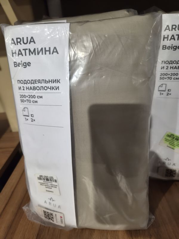 Комплект постельного белья ARUA Натмина бежевого цвета в прозрачной упаковке на деревянной полке: пододеяльник и 2 наволочки 200×200 / 50×70 см, на упаковке наклейка с ценой и QR; справа такая же упаковка с наклейкой, выделенной зелёным маркером