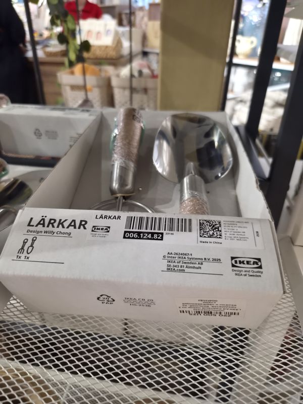 Коробка садового набора IKEA LÄRKAR (рыхлитель и совок) на полке из металлической сетки, на боковой стенке коробки наклейка с ценой Scandi Home; на заднем плане размытые посетители и растения