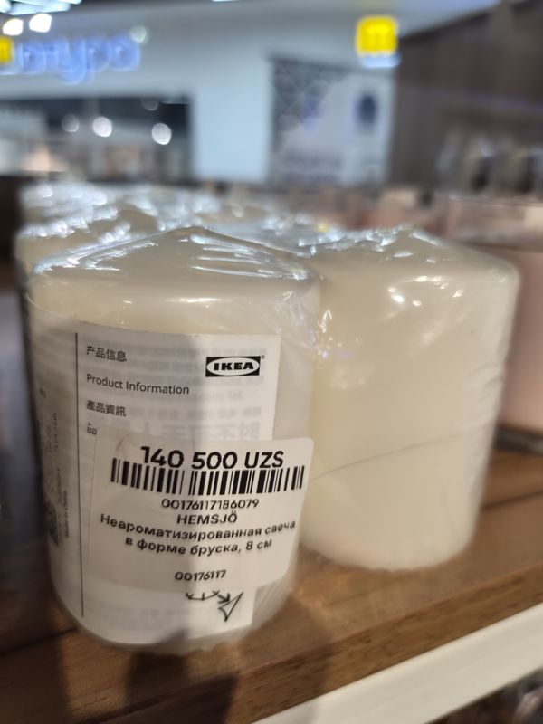 Свечи IKEA HEMSJÖ в форме бруска в плёнке на деревянной полке, на упаковке наклейка с ценой Scandi Home; справа розовые свечи в стакане