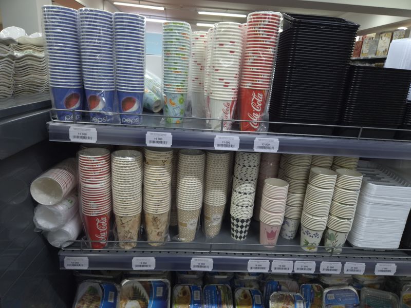 Стеллаж с одноразовыми бумажными стаканами (Pepsi, Coca-Cola, кофейные, цветные), чёрными пластиковыми контейнерами и тарелками, ценники 2 000–21 000