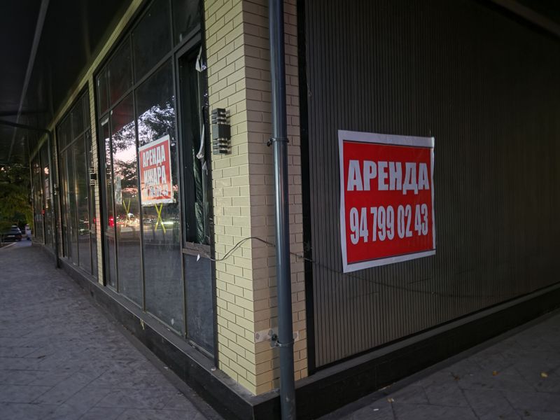 Угол здания с остеклённым свободным помещением на первом этаже: на стене красный плакат «Аренда», в окне такой же плакат «Аренда / Ижара»