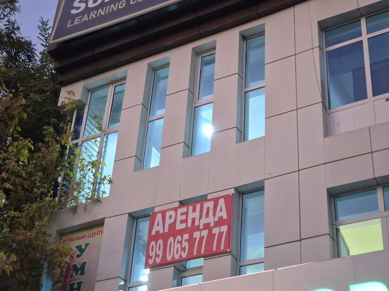 Вечером: красный плакат «Аренда» с номером телефона висит под окном второго этажа светлого здания; на фасаде видны вывеска учебного центра