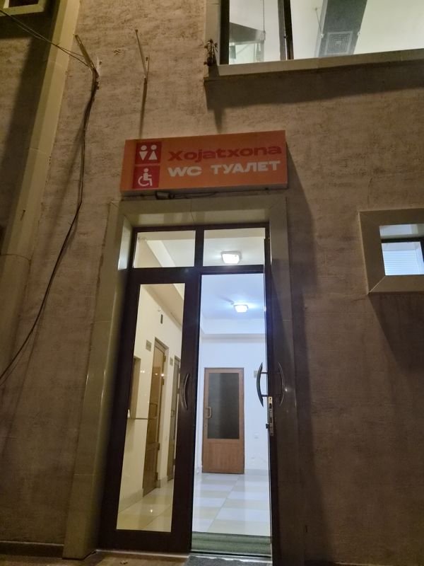 Вход в туалет парка: оранжевая вывеска «Xojatxona / WC Туалет» со значком для инвалидов над стеклянной дверью